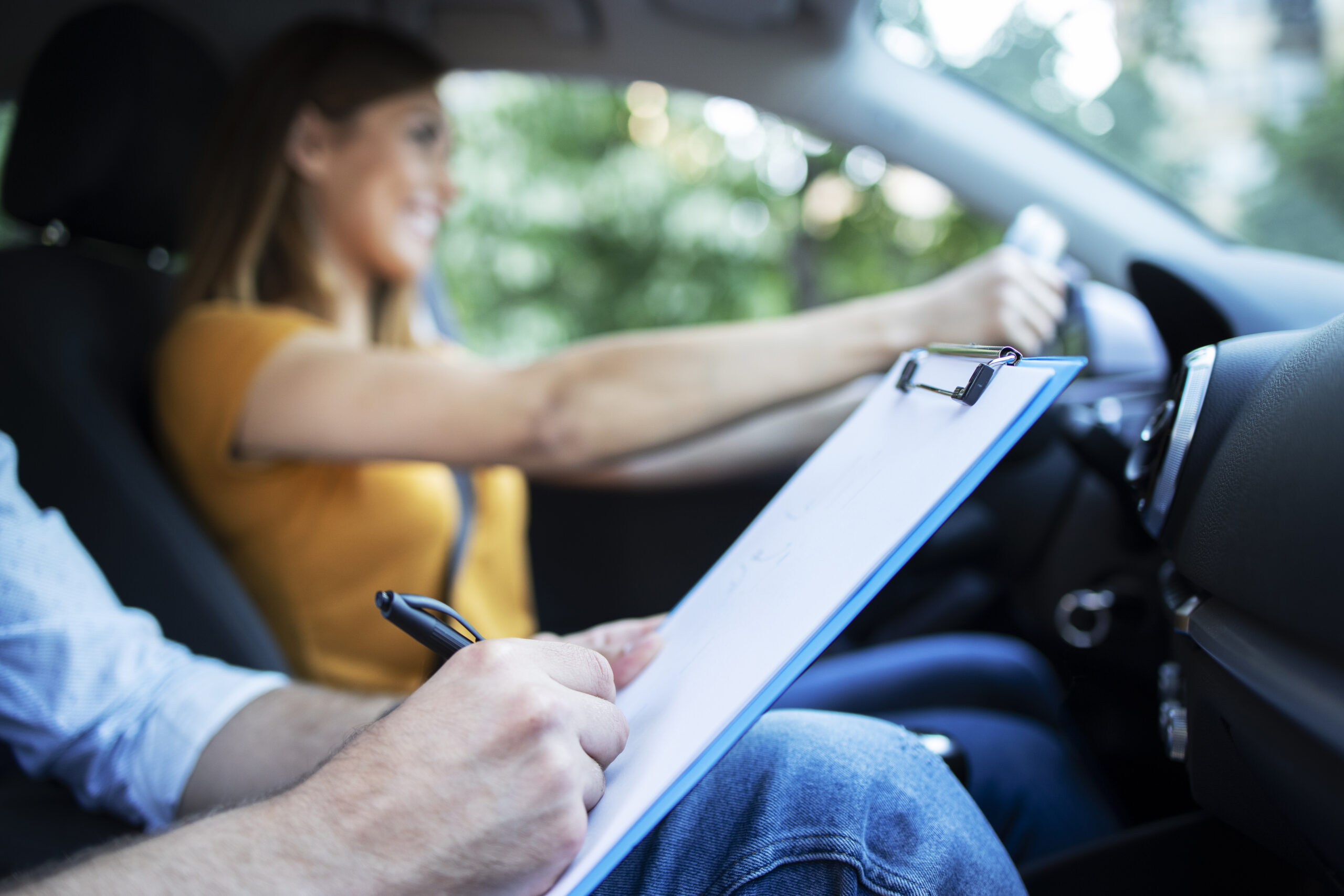 Close up view of driving instructor holding checklist while in background female student steering and driving car.