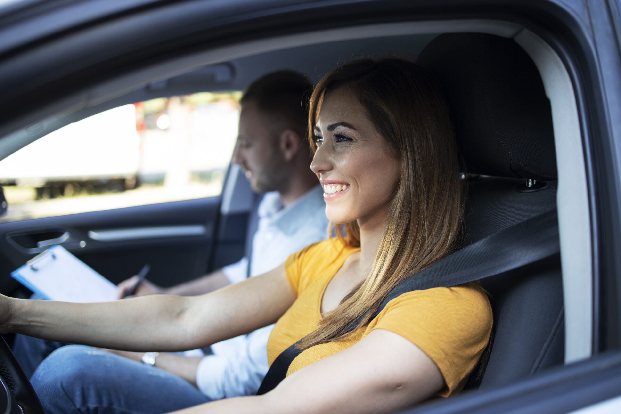 Close up view of female student driving a car and instructor holding checklist in background.